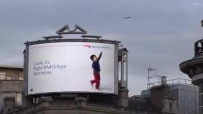 A billboard in London where the kid points at a passing plane and provides the flight info