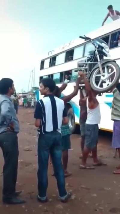 Man carries a motorcycle atop his head while climbing a ladder to place it onto the roof of a bus.