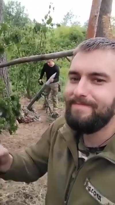 Ukranian soldier enjoying a beverage while his comrade fires a mortar.