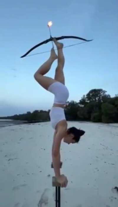 Woman hits a bulls eye with a flaming arrow while doing a headstand using her feet.