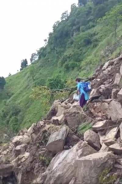 A vaccinator from Nepal ensuring continuity of essential service of immunization despite pandemic and disaster. She is carrying 2 bags, full of vaccines and drops, moving uphill through a route blocked by a recent landslide.