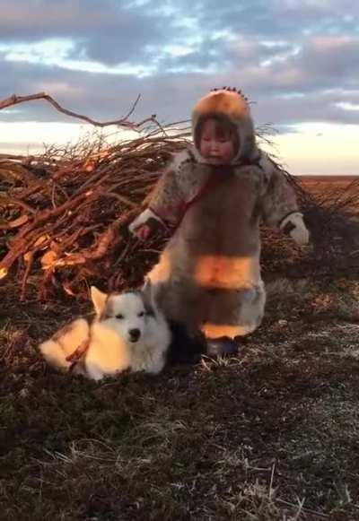 A tiny northern girl tries to play with a puppy. A small bells is sewn on the girl's clothes so that parents can hear where the child is.