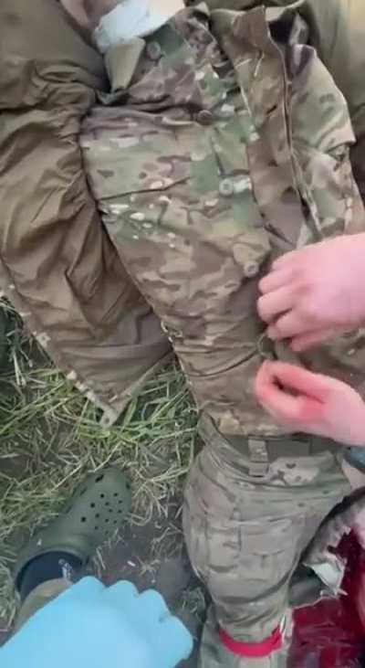 A wounded Russian soldier is lying on the ground already having lost some blood, his comrade cuts open his clothes and injects something in his shoulder area