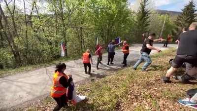 Drunk spectators wrestle in the middle of the road seconds before one of rally cars pass by at high speed