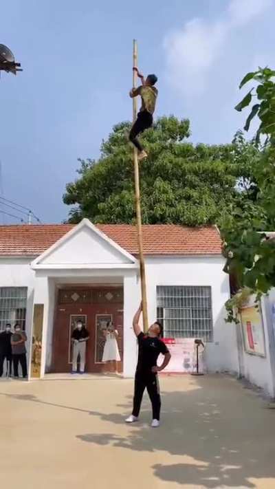 The man is balancing a long bamboo stick on his shoulder. Second dude climbs up the stick quickly and does acrobatics on it.