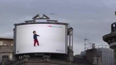 A British airline billboard in London where a kid points at a passing plane and gives you the information of that flight