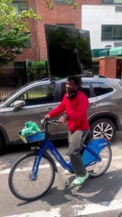 Dude balances a 45 inch TV on his head while riding a bicycle