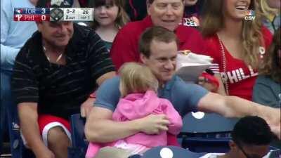 A dad grabs a foul ball while holding his child in a baseball stadium.